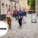 Menschen gehen auf einem Kopfsteinpflasterweg, einige halten Konferenzmaterialien in den Händen, im Hintergrund sind Gebäude zu sehen.