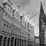 Schwarzweißfoto, das den historischen Charme der Altstadt von Münster einfängt, mit einem hohen Kirchturm, der in den wolkigen Himmel ragt – eine perfekte Szene für eine GEO EPOCHE oder Historische Tour.