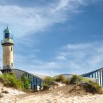 Leuchtturm mit modernem Gebäude an einem Sandstrand unter einem blauen Himmel mit Wolken.