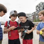 Vier Jungen mit Rucksäcken und Wasserflaschen versammeln sich um ein Tablet und erkunden die Details einer Stadtrallye im pulsierenden Stadtbild von Düsseldorf.