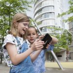 Lächelnd erkunden zwei Kinder im Freien, umgeben von der üppigen Vegetation des Düsseldorfer Nordparks, ein Tablet. Zwischen den Bäumen lugen moderne Gebäude hervor, die den Rahmen für ihr spannendes GEOLINO-Stadtrallye-Abenteuer bilden.