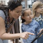 Umgeben von der malerischen Natur des Düsseldorfer Nordparks sind eine Frau und zwei Kinder auf ihrem Tablet in eine GEOLINO-Stadtrallye vertieft.