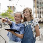 Vor den modernen Gebäuden Düsseldorfs stehen zwei Kinder bei einer GEOLINO-Stadtrallye im Freien, eines zeigt lächelnd nach vorne, das andere hält ein Tablet in der Hand.