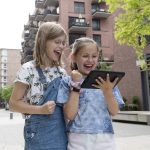 Zwei aufgeregte Kinder nehmen an einer spannenden Stadtrallye teil und starren gemeinsam gespannt auf ein Tablet in einer belebten Stadtstraße. Sie tauchen in die abenteuerlichen Hinweise von GEOLINO ein und planen möglicherweise ihren nächsten Schritt in der Nähe des Düsseldorfer Nordparks.