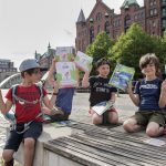 Drei Kinder sitzen draußen im Düsseldorfer Nordpark, halten bunte GEOLINO-Bücher hoch und lächeln, im Hintergrund sind historische Gebäude und Bäume zu sehen.