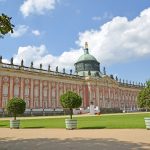 Historischer Palast mit verzierter Fassade, Kuppel und Statuen unter einem blauen Himmel mit flauschigen Wolken. Umgeben von gepflegtem Rasen.