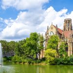 Historische Kirche an einem See mit einem Brunnen, umgeben von Bäumen unter einem teilweise bewölkten blauen Himmel.