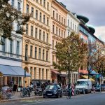 Straße mit bunten Gebäuden, geparkten Autos, Fahrrädern und Fußgängern an einem bewölkten Tag.