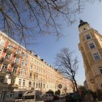 Stadtstraße mit farbenfrohen historischen Gebäuden, blattlosen Bäumen und geparkten Autos unter einem klaren blauen Himmel.