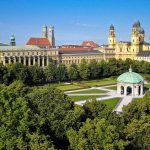 Blick auf den Hofgarten in München mit einem Pavillon, umgeben von Bäumen und Stadtgebäuden unter einem klaren blauen Himmel.