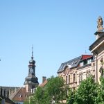 Historische europäische Straße mit reich verzierten Gebäuden, einem Kirchturm und grünen Bäumen unter einem klaren blauen Himmel.