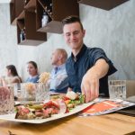 Ein lächelnder Mann greift in einem gemütlichen Restaurant nach Essen auf einem Tablett, im Hintergrund sind andere Gäste.