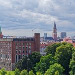 Stadtbild mit Backsteingebäuden, grünen Bäumen und zwei markanten Kirchtürmen unter einem bewölkten Himmel.