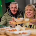 Ein Mann und eine Frau lächeln, während sie in gemütlicher Atmosphäre Essen auf Holztabletts servieren.