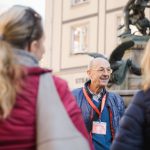 Ein älterer Mann mit einem roten Schlüsselband führt eine Reisegruppe nach draußen in die Nähe einer Statue und lässt sie in die reiche Geschichte der Inneren Neustadt Dresdens eintauchen.