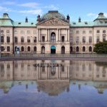 Historisches Gebäude mit grünen Dächern, die sich an einem klaren Tag in einem großen, stillen Teich spiegeln.