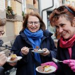 Drei Frauen genießen lächelnd und plaudern im Freien ein Dessert und tragen Winterjacken und Schals.