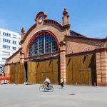 Ein Radfahrer und Fußgänger in der Nähe eines historischen Backsteingebäudes mit großen braunen Türen und einer Uhr unter einem klaren blauen Himmel.