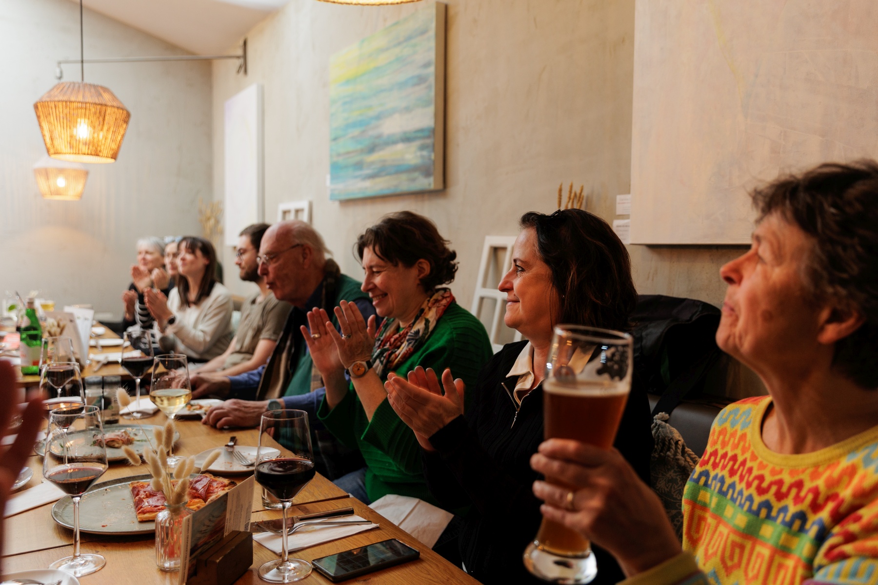 In einem gemütlichen, warm beleuchteten Raum in Frankfurt sitzen Menschen an einem Esstisch, klatschen und halten Getränke in der Hand, während sie einen herrlichen Dinner-Hopping-Abend genießen.