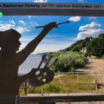 Silhouette eines Malers vor der Kulisse eines sonnigen Flussufers mit Menschen, die am Sandstrand entspannen. Die Szene fängt die Essenz der schönen Orte entlang der malerischen Ufer des Hamburger Wassers ein.