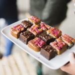 Ein Tablett mit verschiedenen Schokoladen- und Vanillekuchen, jeder mit rosa Zuckerguss gekrönt, wird von einer Person gehalten, die eine kulinarische Reise genießt, die einer süßen Stadtführung gleicht.